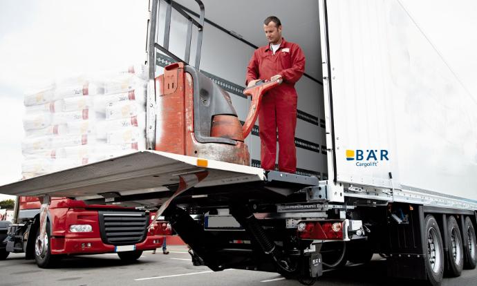 Chauffeur met elektrische palletwagen en zware pallet op het platform van een onderrijdbare Bar-hefbrug
